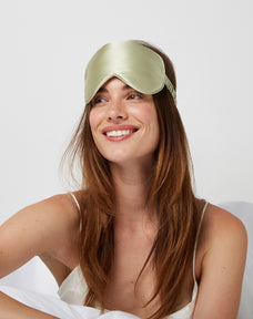 Woman looking happy while sitting comfortably in bed wearing a Luxury Soft Silk Sleeping Eye Mask from Dore and Rose in the color Olive Green