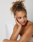 Woman looking happy while sitting comfortably in bed wearing a Luxury Soft Silk scrunchie from Dore and Rose in the color Gray