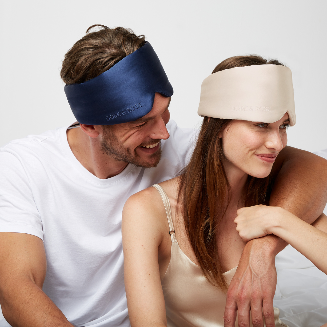 Man and woman both smiling and wearing silk sleeping eye masks in navy blue and in champagne