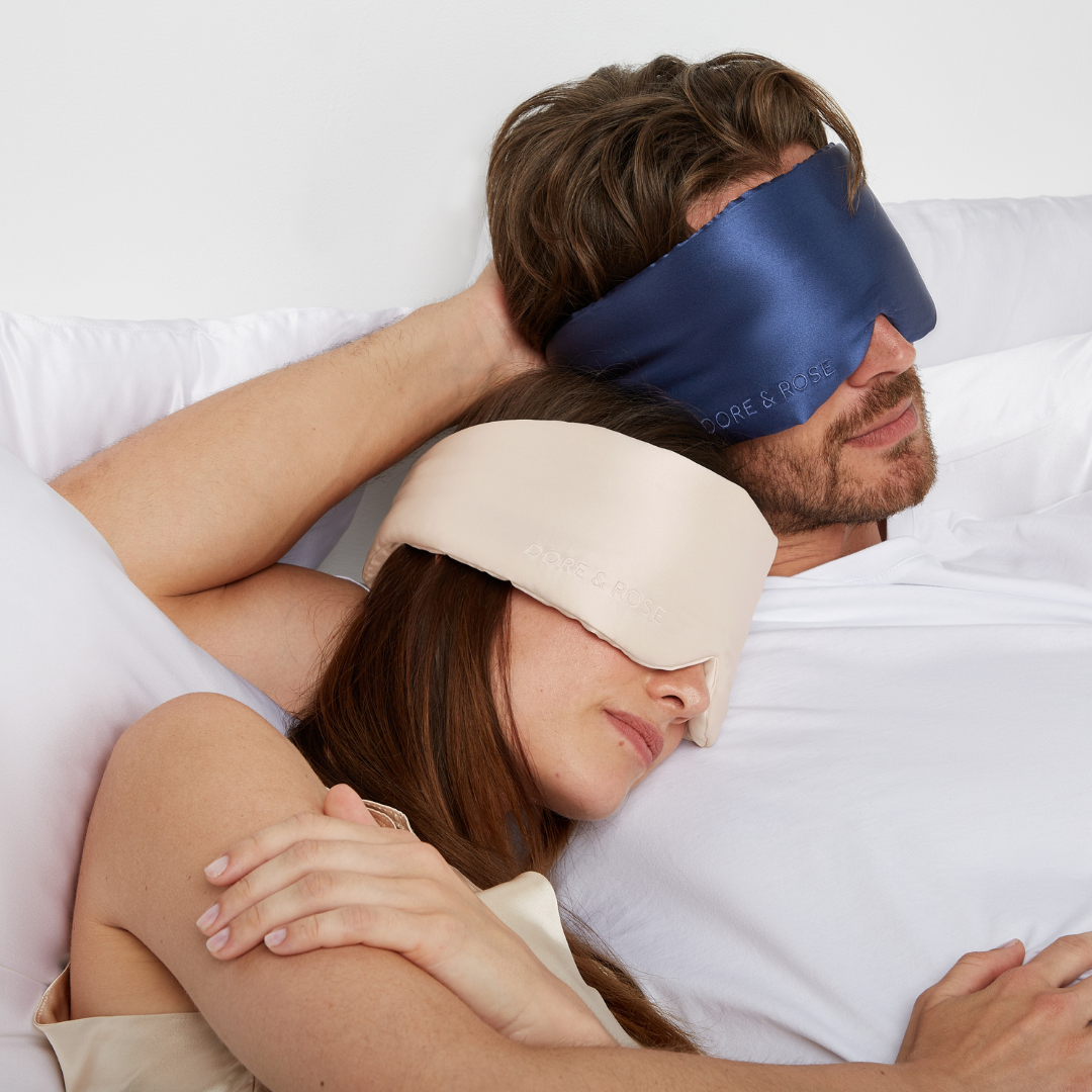 Man and woman lying next to each other both wearing silk sleeping eye masks in navy blue and in champagne