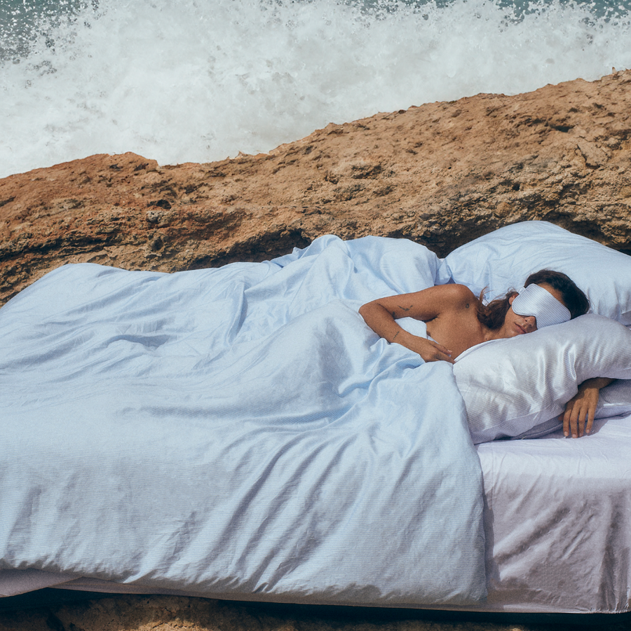 Woman sleeping in bed outdoors in silk pillowcases wearing a Premium Stretchable Silk Sleep Eye Mask from Dore and Rose in the color Blue and White stripes