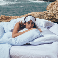 Woman lying comfortably in bed outdoors in silk pillowcases wearing a Luxury Soft Silk Sleeping Eye Mask from Dore and Rose in the color Blue and White stripes