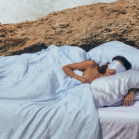 Woman sleeping in bed outdoors in silk pillowcases wearing a Premium Stretchable Silk Sleep Eye Mask from Dore and Rose in the color Blue and White stripes