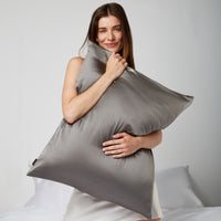 Woman sitting on a white bed while hugging a pillow with a gray silk pillowcase