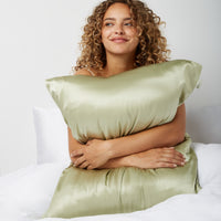Woman sitting in bed looking happy hugging a pillow with a silk pillowcase from Dore and Rose in the color Olive Green