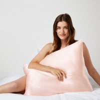 Woman sitting comfortably in bed hugging a pillow with a silk pillowcase from Dore and Rose in the color Pink