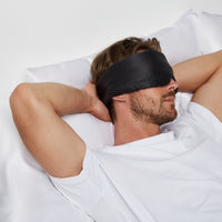 Man lying comfortably in bed wearing a Silk Sleeping Eyemask from Dore and Rose in the color Black