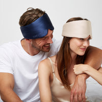 Happy Couple sitting in bed wearing a Silk Sleeping Eyemask from Dore and Rose in the colors Champagne Beige for the woman and Navy Blue for the Man