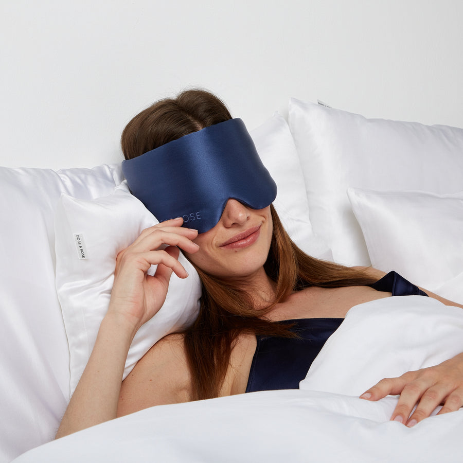 Woman lying comfortably in bed wearing a Silk Sleeping Eyemask from Dore and Rose in the color Navy Blue