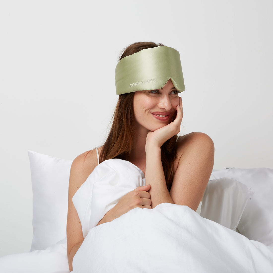 Woman smiling while sitting in bed wearing a Luxury Soft Silk Sleeping Eye Mask from Dore and Rose in the color Olive Green