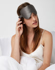 Woman sitting comfortably in bed wearing a Luxury Soft Silk Sleeping Eye Mask from Dore and Rose in the color Gray