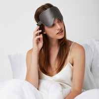 Woman sitting comfortably in bed wearing a Luxury Soft Silk Sleeping Eye Mask from Dore and Rose in the color Gray