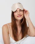 Woman just woke up sitting comfortably in bed wearing a Luxury Soft Silk Sleeping Eye Mask from Dore and Rose in the color Champagne Beige