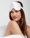 Woman looking happy while sitting comfortably in bed wearing a Luxury Soft Silk Sleeping Eye Mask from Dore and Rose in the color White