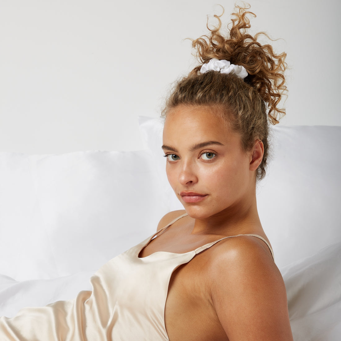 Woman looking happy while sitting comfortably in bed wearing a Luxury Soft Silk Scrunchie from Dore and Rose in the color White